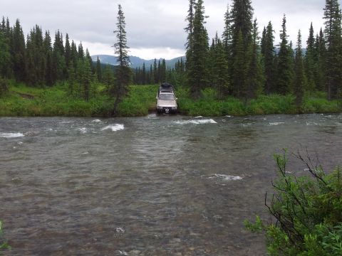 1990 Chevrolet Suburban - A tan SUV navigates a shallow river surrounded by dense pine forest and distant mountains.