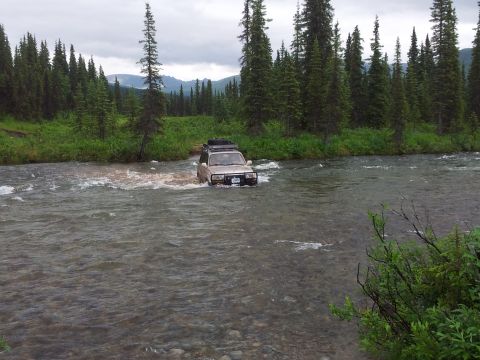 1990 Chevrolet Suburban - A tan SUV navigates a shallow river surrounded by dense forest and distant mountains under an overcast sky.