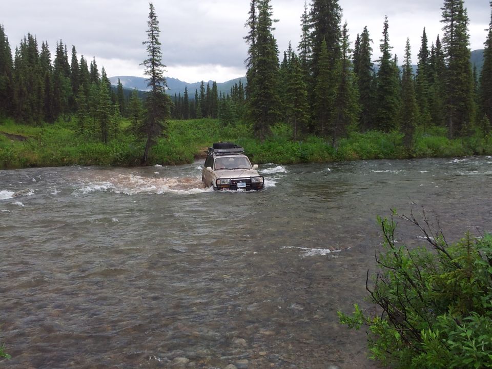 Tan SUV driving through a river with trees and mountains in the background