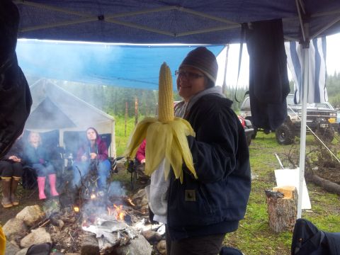 1990 Chevrolet Suburban - A person proudly displays a giant ear of corn at an outdoor gathering with a campfire and tents in the background.
