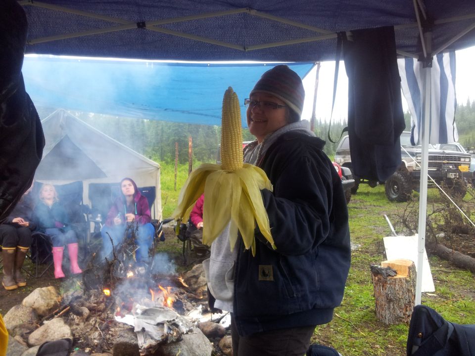 Person holding a large ear of corn at a campsite with a campfire and tents