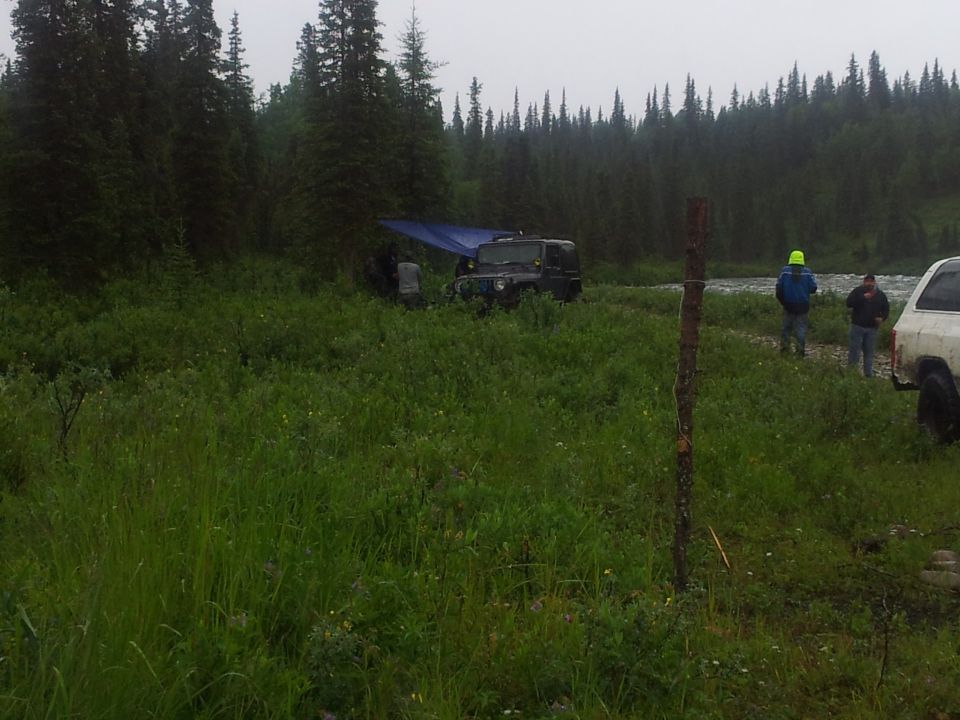 Two SUVs parked in a grassy area near a river, with a blue tarp shelter set up