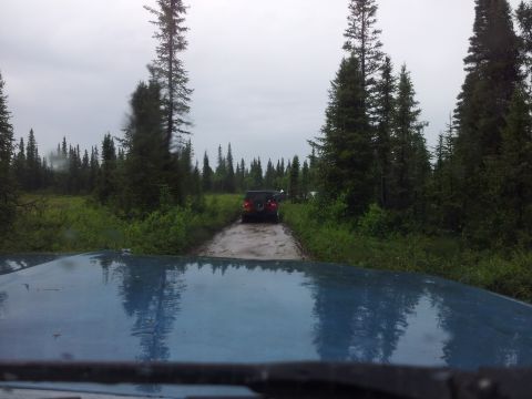 1990 Chevrolet Suburban - Two SUVs navigate a wet, muddy road through a dense forest under an overcast sky.
