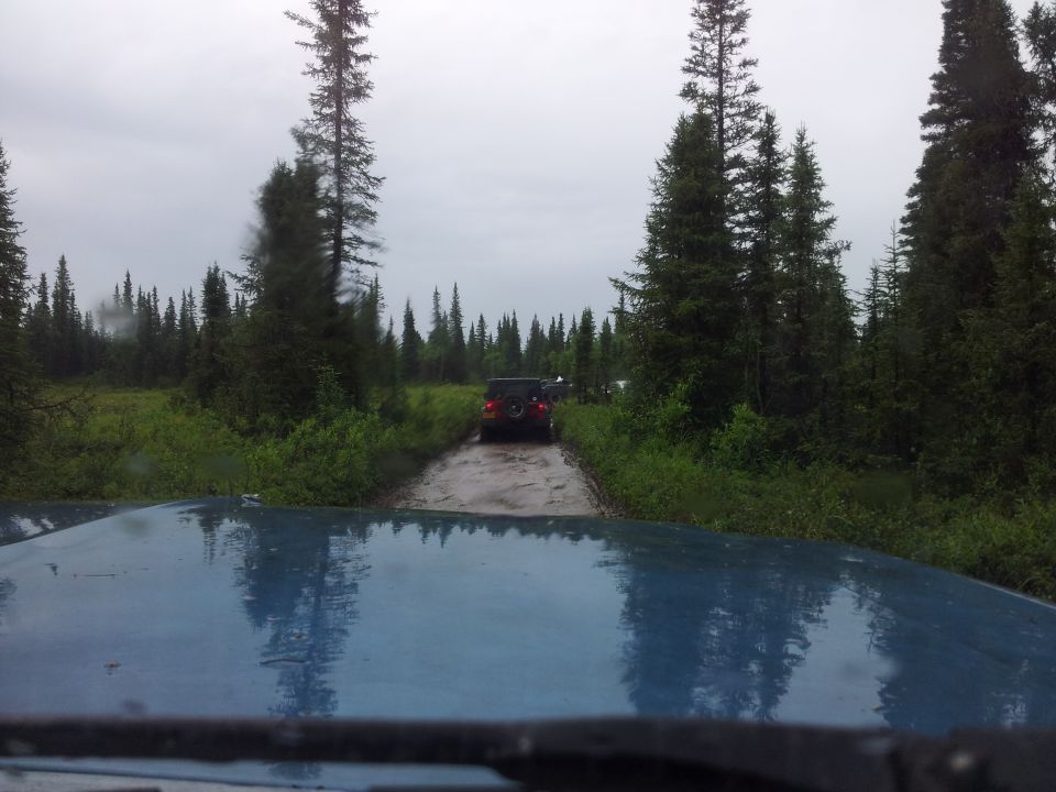 Blue vehicle hood reflects trees and sky, with two SUVs driving on a muddy forest road ahead