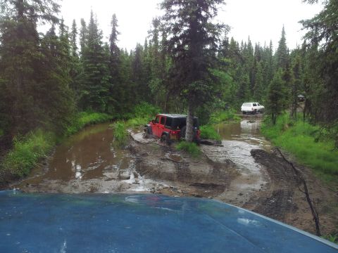 1990 Chevrolet Suburban - A red Jeep Wrangler is stuck in a muddy water crossing on a remote forest road, with a white SUV following behind.