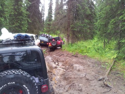 1990 Chevrolet Suburban - Multiple off-road vehicles are stuck in deep mud on a trail surrounded by dense forest.