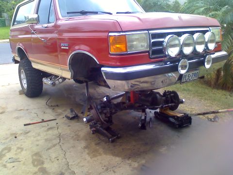 1990 Ford Bronco - A red 1990 Ford Bronco is shown with its front end disassembled, supported by jack stands.