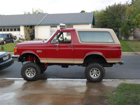 1990 Ford Bronco - This lifted red Ford Bronco is parked on a residential street, showing off its off-road modifications.