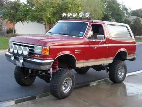 1990 Ford Bronco - A lifted 1990 Ford Bronco with off-road lights and a two-tone paint scheme is parked on a wet street.