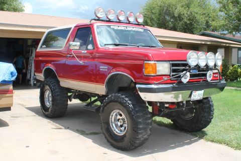 1990 Ford Bronco - Lifted red Ford full-size Bronco on large mud-terrain tires with chrome wheels, roof light bar and front brush guard, parked in a driveway.