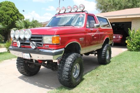 1990 Ford Bronco - Red lifted late-1980s Ford Bronco with roof-mounted light bar, brush guard lights, and large mud-terrain tires parked in a driveway.