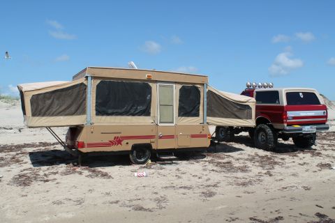 1990 Ford Bronco - A red and white 1990 Ford Bronco tows a tan pop-up camper across a sandy beach under a clear blue sky.