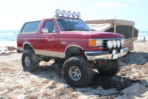 1990 Ford Bronco - Lifted 1990 Ford Bronco parked on a sandy beach with a pop-up camper in the background.