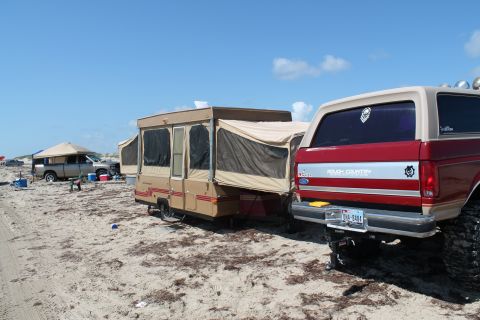 1990 Ford Bronco - A red 1990 Ford Bronco is parked on a sandy beach next to a pop-up camper, with other vehicles and tents visible in the background.