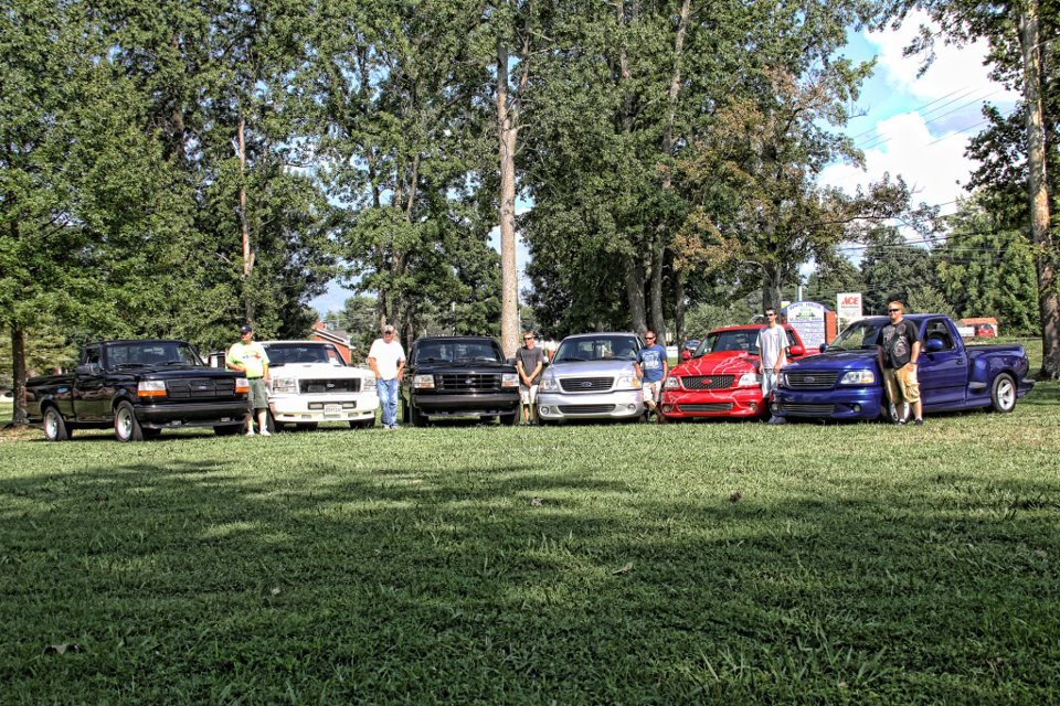 Several Ford F-150 SVT Lightning trucks parked on grass, with trees and a building in the background