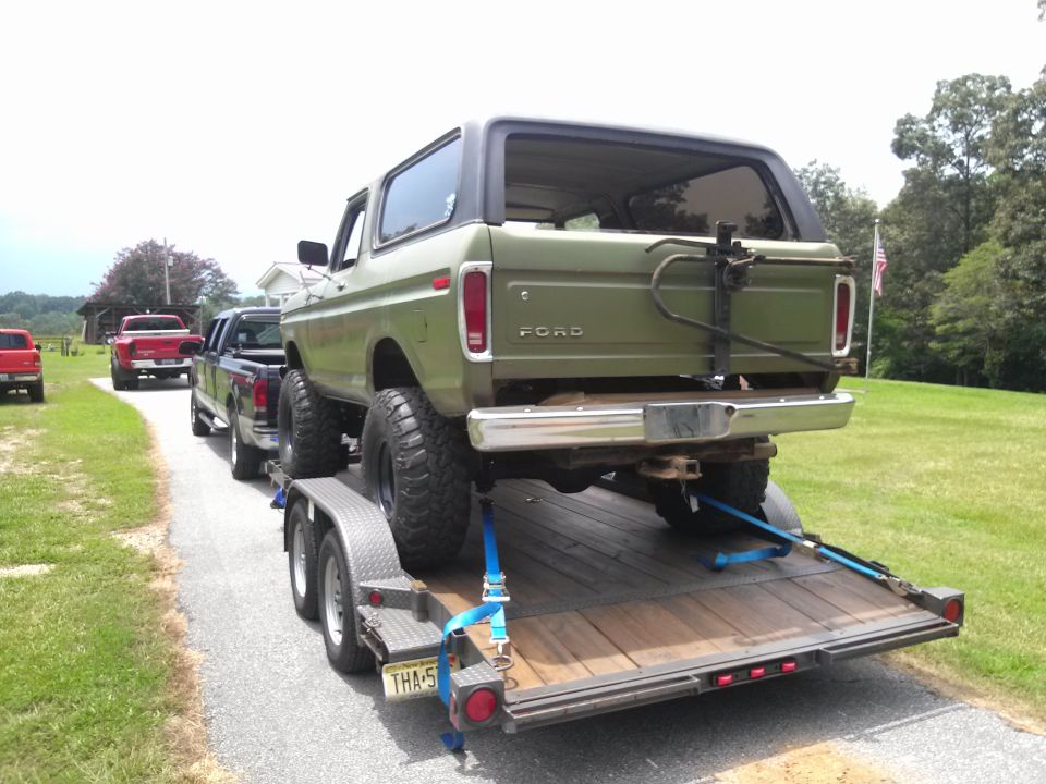 Green Ford Bronco on a trailer, with two pickup trucks behind it on a road