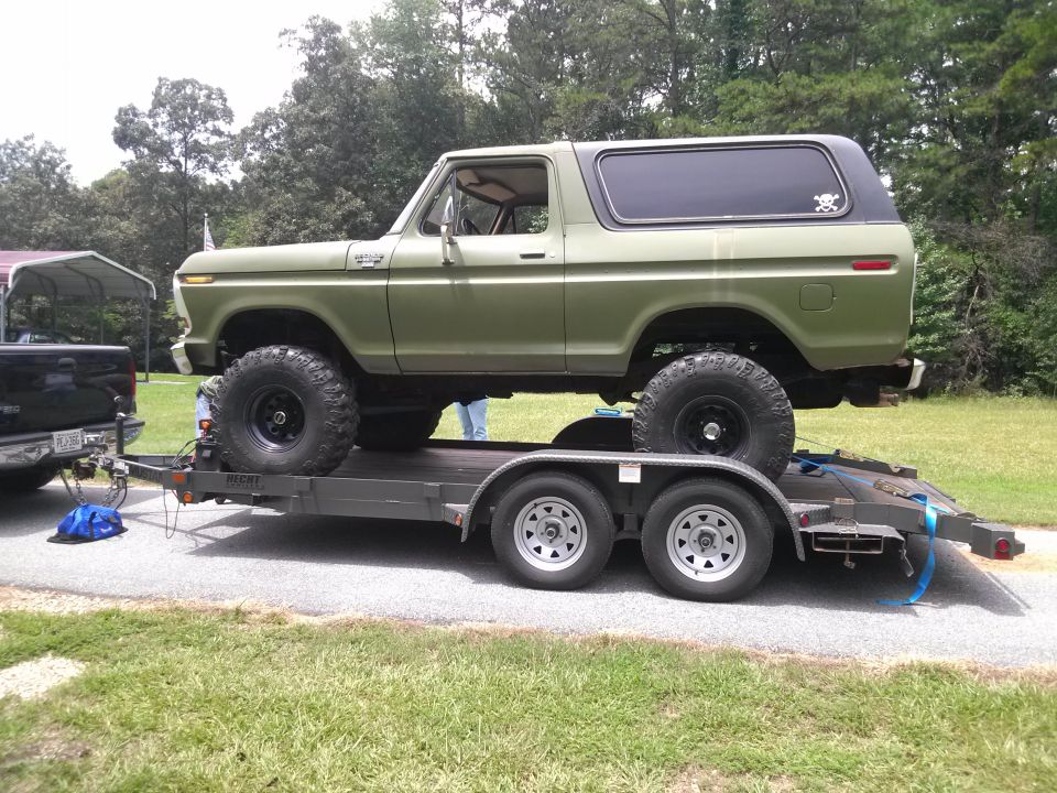 Olive green Ford Bronco with lifted suspension and large tires on a trailer, being towed by a black pickup truck
