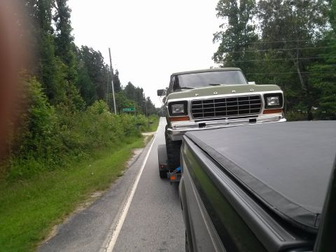 1979 Ford Bronco - A green Ford truck is being towed on a trailer down a two-lane road with trees on the side.