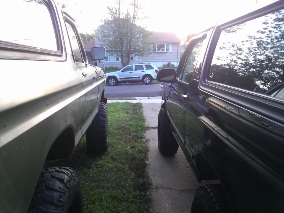 Two lifted SUVs, one light-colored and one dark, parked on a driveway with a house and another SUV in the background