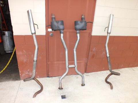 2003 Ford Crown Victoria - pair of fabricated dual exhaust systems with mufflers and tailpipes leaning upright against a painted concrete block wall.