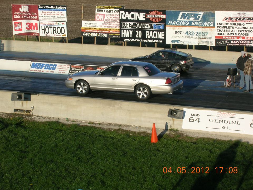 Silver Ford Crown Victoria racing another car on a drag strip. Signs for various sponsors are visible in the background