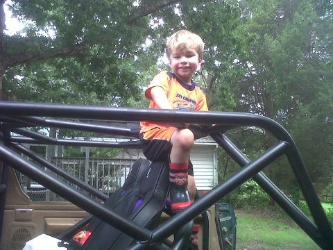 1990 Ford Bronco - A young boy smiles while sitting atop a custom roll cage, with trees and a house in the background.
