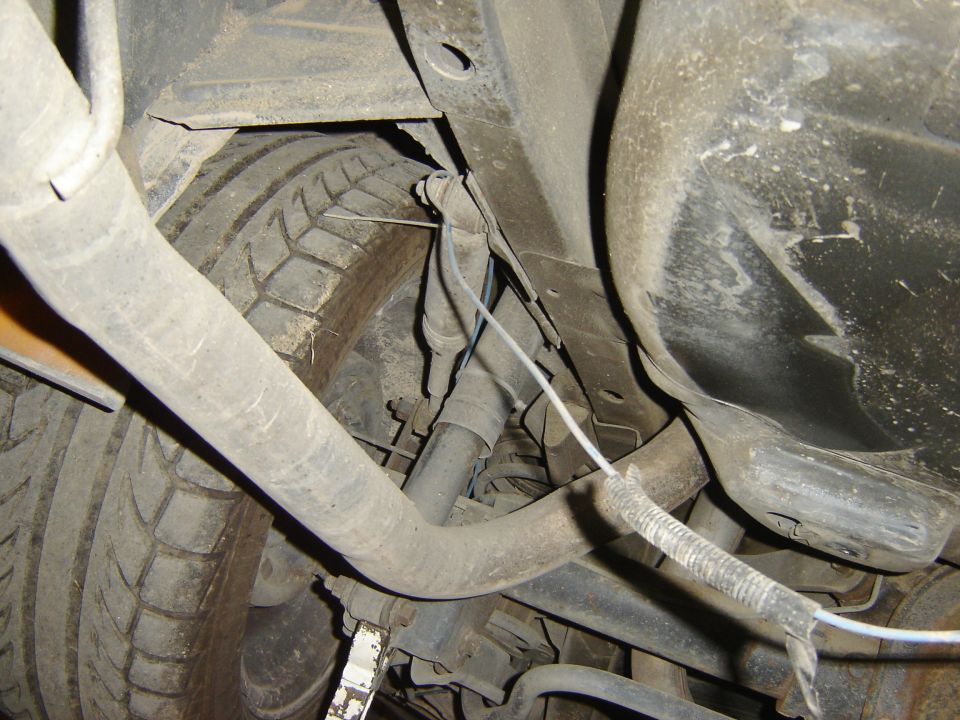 Underside view of a car's rear tire and suspension components. Shows exhaust pipe and wiring