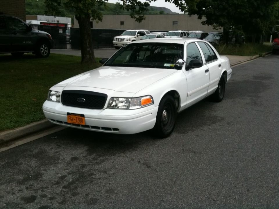 White sedan parked on street, black grille, orange turn signals, steel wheels. Other cars and buildings in background