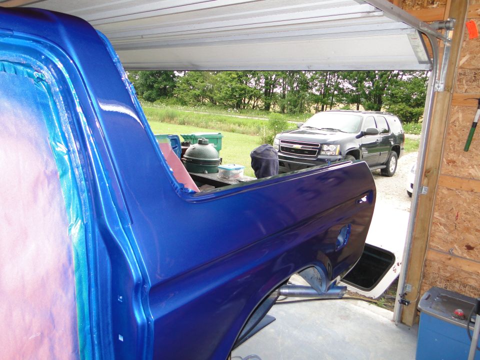 Side view of a blue vehicle body with fresh paint, parked in a garage with another SUV outside