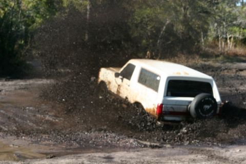1988 Ford Bronco - A white 1988 Ford Bronco is shown struggling through a muddy off-road trail, kicking up a large spray.