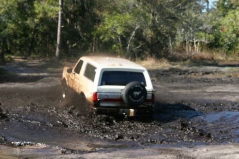 1988 Ford Bronco - A white SUV navigates through a muddy terrain, with trees visible in the background.
