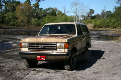 1988 Ford Bronco - A tan 1987 Ford Bronco is parked on a dirt road with trees in the background.
