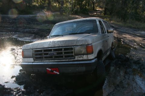 1988 Ford Bronco - A dusty Ford Bronco navigates a muddy trail, kicking up water.