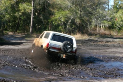 1988 Ford Bronco - A white 1987 Ford Bronco navigates a muddy off-road course, kicking up water and dirt.