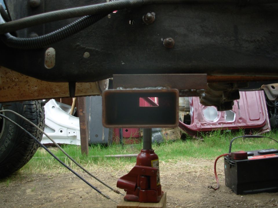 Vehicle frame underside with red bottle jack holding a rusty crossmember, battery nearby