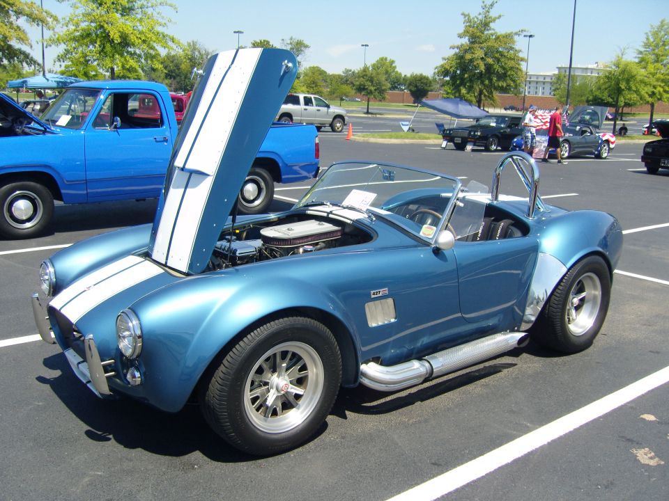 Blue AC Cobra replica, hood open, showing engine. Another blue pickup truck is parked to the left