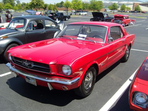 2007 Ford Mustang - A bright red 1965 Ford Mustang is parked in a lot next to a grey Volkswagen Beetle.