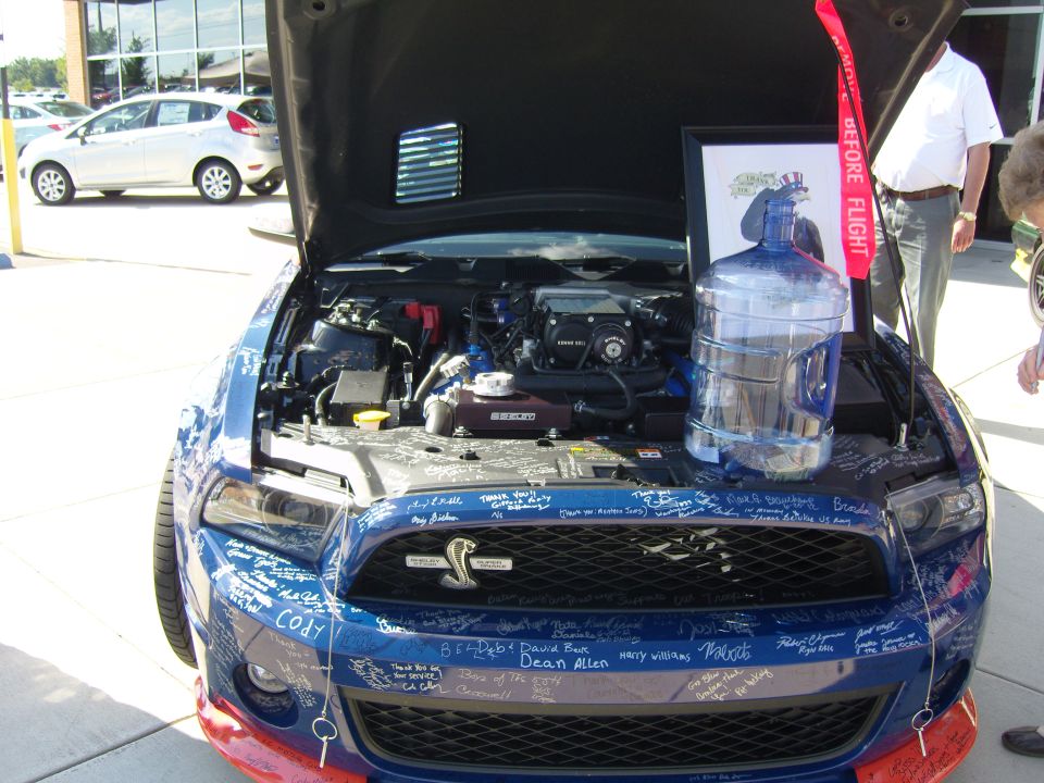 Blue Ford Mustang with hood open, engine visible, covered in signatures. A framed picture and water jug sit on top of the engine