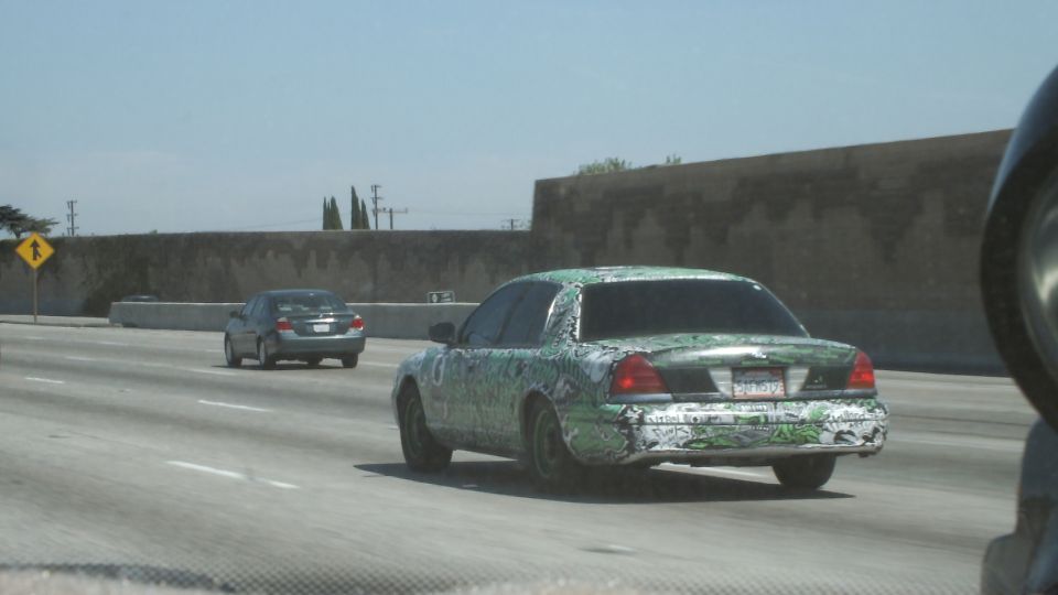 Green graffiti-wrapped full-size sedan driving on a freeway past a concrete wall