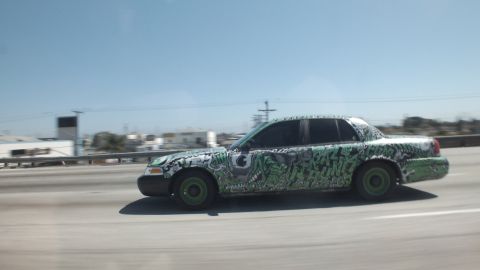 2003 Ford Crown Victoria - Side view of a green-and-black art-wrapped sedan moving along a highway near a city