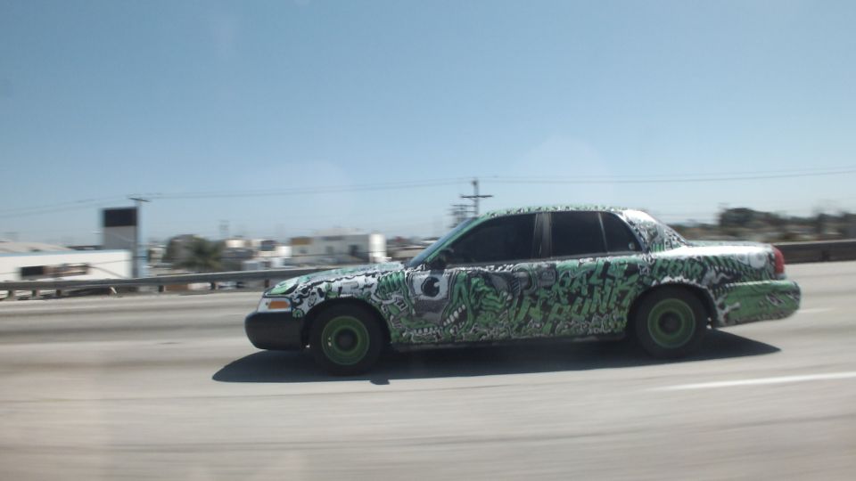 Graffiti-wrapped sedan driving along an elevated freeway with city buildings behind