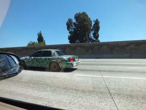 2003 Ford Crown Victoria - Art-wrapped sedan cruising the highway with a sound wall and trees in the background