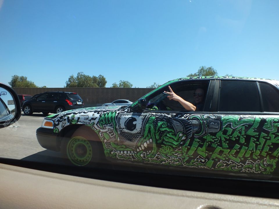Driver flashing peace sign from a green graffiti-wrapped sedan in freeway traffic