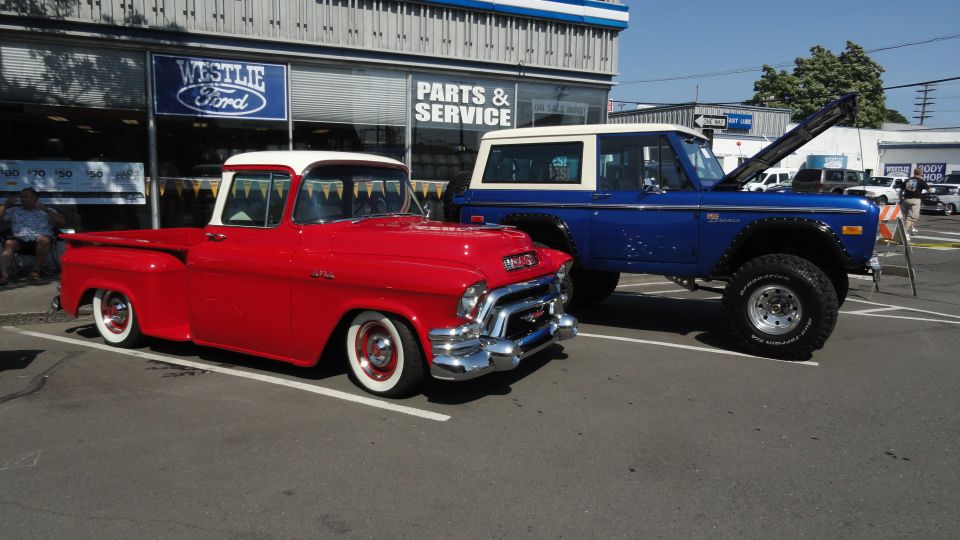 Red 1950s GMC pickup truck parked next to a blue Ford Bronco with hood open. Westlie Ford dealership in background