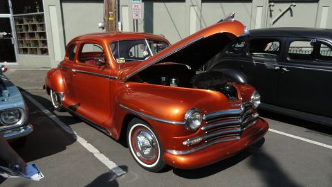 1964 Ford Fairlane - A shiny orange coupe hot rod is parked with its hood up, next to a dark sedan.