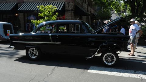 1964 Ford Fairlane - A black 1955 Chevrolet sedan is parked on a street with its hood open, while onlookers gather around.