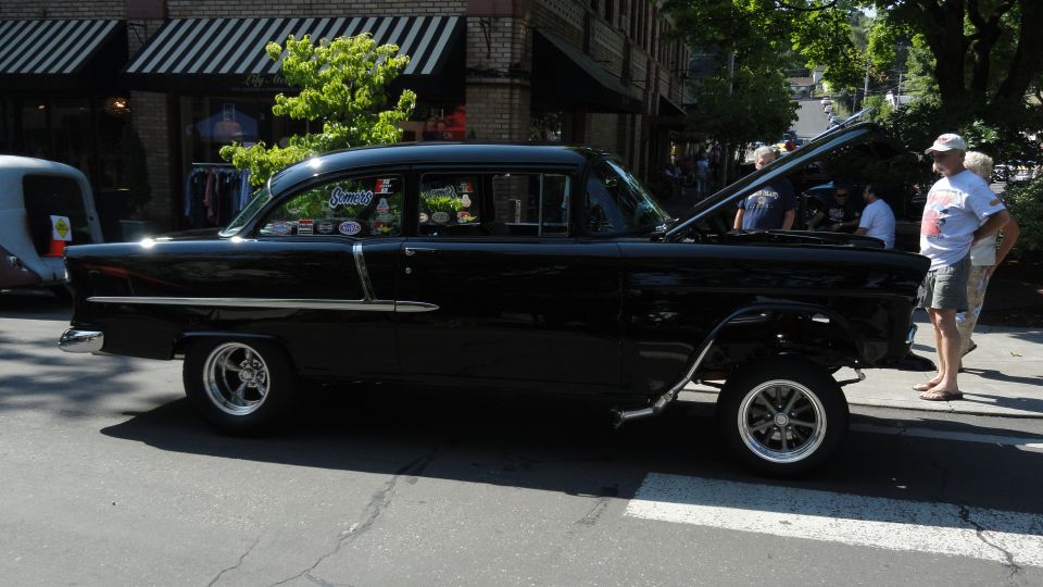 Black 1955 Chevrolet sedan, side view, hood open, chrome trim, custom wheels, people standing nearby