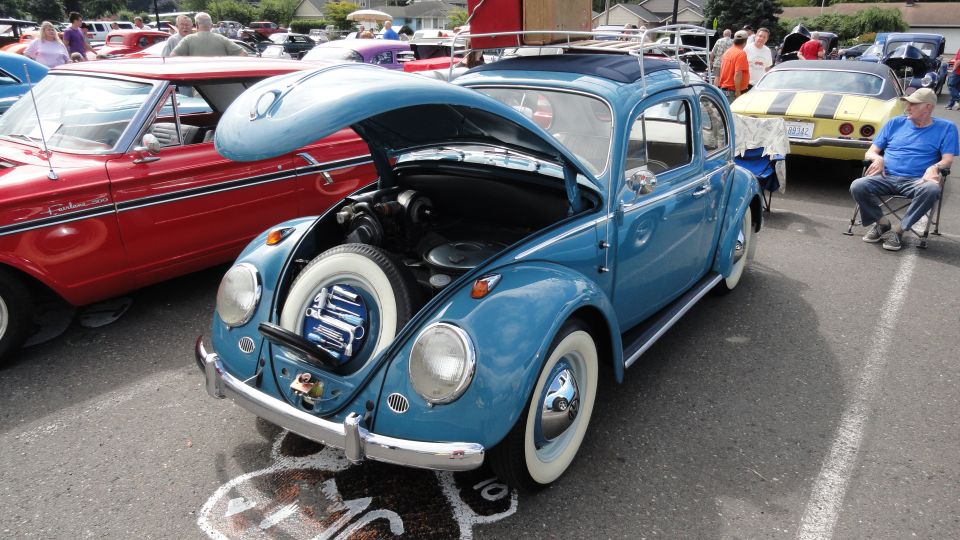 Blue VW Beetle with hood open, showing spare tire. Red car next to it. Yellow car and man in chair in background