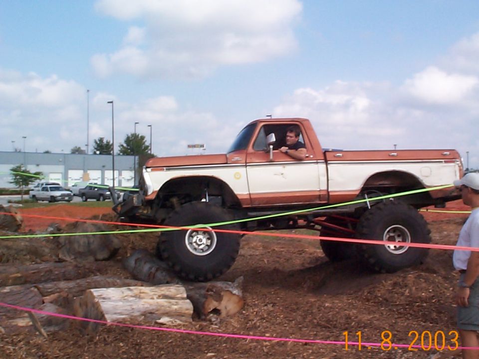 Lifted orange and white 1970s Ford pickup on huge mud tires crawling over logs