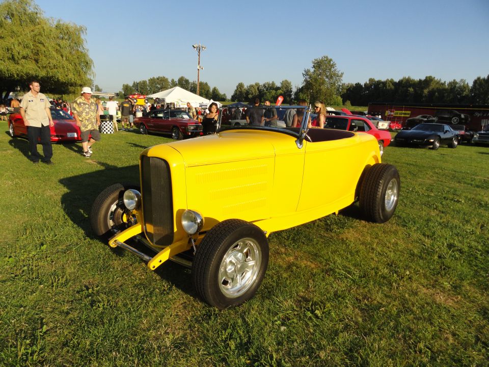 Yellow hot rod convertible with wide rear tires parked on grass at an outdoor car show. Other cars and people are visible in the background